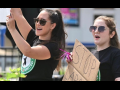 Two young women in Starbucks Workers United T-shirts stand outdoors, holding up cardboard signs, mouths open to yell or chant.