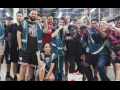 A group of workers, diverse in race and gender, poses together on the factory floor. Most appear young; one wears a prosthetic arm. A few have fists raised and most smile or wear determined expressions. Most wear shorts and some wear blue work vests. 