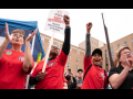 Joyful people in red shirts throw their fists in the air. One has a "CWA On Strike Against Mercy Hospital of Buffalo" picket sign. Outdoors.