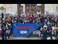 Large, multiracial, masked crowd stands on the steps of a public buliding. Many have fists in the air. Back row holds ATU banners. Front row holds a giant banner that reads "Hazard pay now!" with a line drawing of a bus.