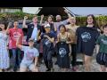 Eleven smiling people pose outside. Some are holding up silkscreened T-shirts with Tender Claws union logo showing a claw. Many are raising one fist, and one is pointing at another person in the group.