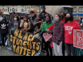 A multiracial and multigenerational crowd rallies outside. Several help hold a large banner: "BLACK STUDENTS MATTER." Others hold signs: "WE WON'T DIE FOR THE DOE" and one wears a MORE T-shirt.