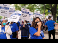 People in blue "INA" shirts march. Some carry picket signs "Safety in Numbers," "Respect Nurses!" "Unfair Labor Practice Strike." One speaks into a bullhorn mic.