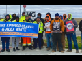 16 people stand facing the camera. Most are wearing jeans and neon yellow or orange jackets or hoodies. Several are helping to hold a blue banner: "Rehire Edward Clarke and Alex White, no retaliation or favoritism." Others hold signs: "180 hours paid time off, unionize KCVG" and "Unionize Amazon."