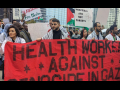 A group of people in white medical coats stand behind a red banner reading ‘Health Workers Against Genocide in Gaza.’