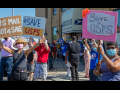 A group of people, masked, socially distanced, and diverse in race and gender, stand outside the Old Bedford Station post office on a sunny day. They hold up handmade signs: "US Mail Not 4 Sale" and "Save USPS." Someone in the back has a camera set up on a tripod. Similar printed signs from APWU are also visible.