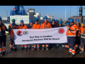 Fourteen workers in orange vests, blue hardhats, and workboots stand in a port in bright sunlight. They hold a big white banner that says "One way or another, European dockers will be heard" with logos of the SDU and the European Dockworkers Council. Most but not all appear to be white men.