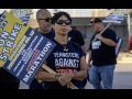 A woman with a strike picket sign and a ‘Teamsters Against Trump’ T-shirt looks past the camera