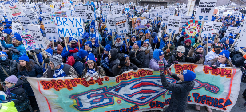 a crowd of educators wearing coats and matching blue MFT knit hats hold picket signs and a banner that reads "districts refuse to pay for ESPs, our children need heroes."