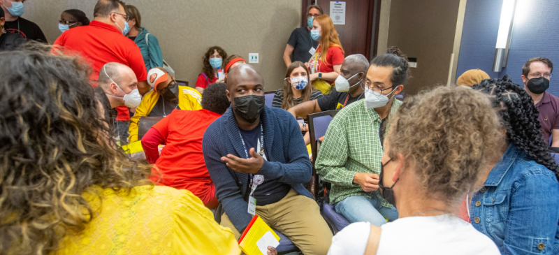 Masked people sit in a circle indoors, talking, in a hotel conference room at the 2022 Labor Notes Conference