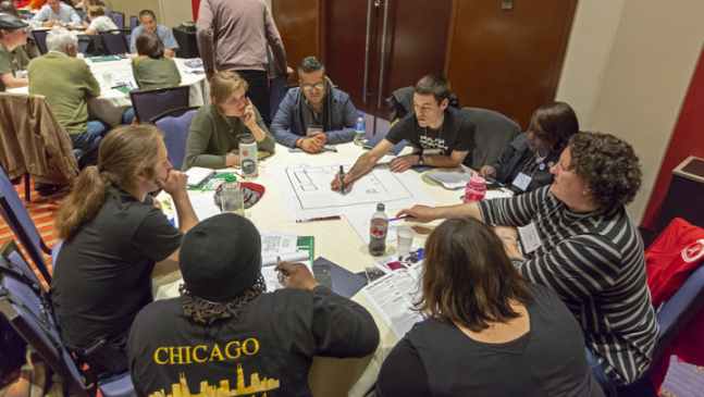 A group of 8 people sit at a round table, as one person draws a large diagram on paper on the table