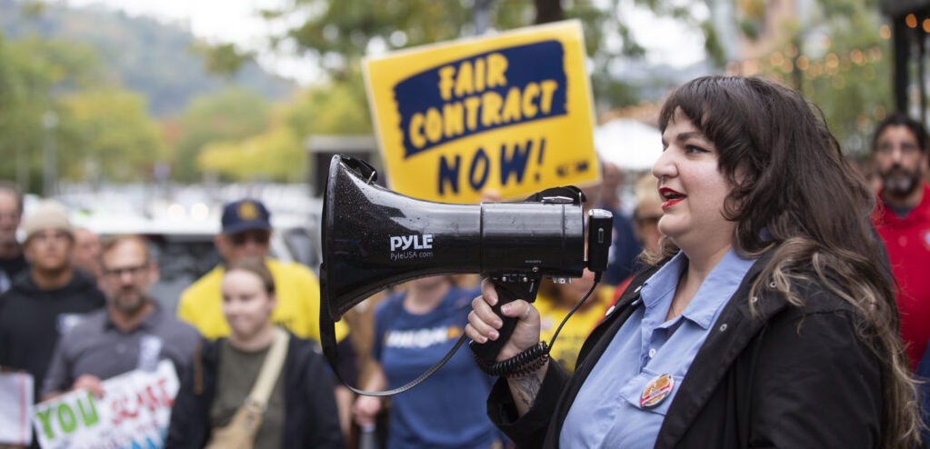 A woman with long dark hair speaks into a megaphone to a crowd that is visible beyond her, one holding a “Fair Contract Now” sign.