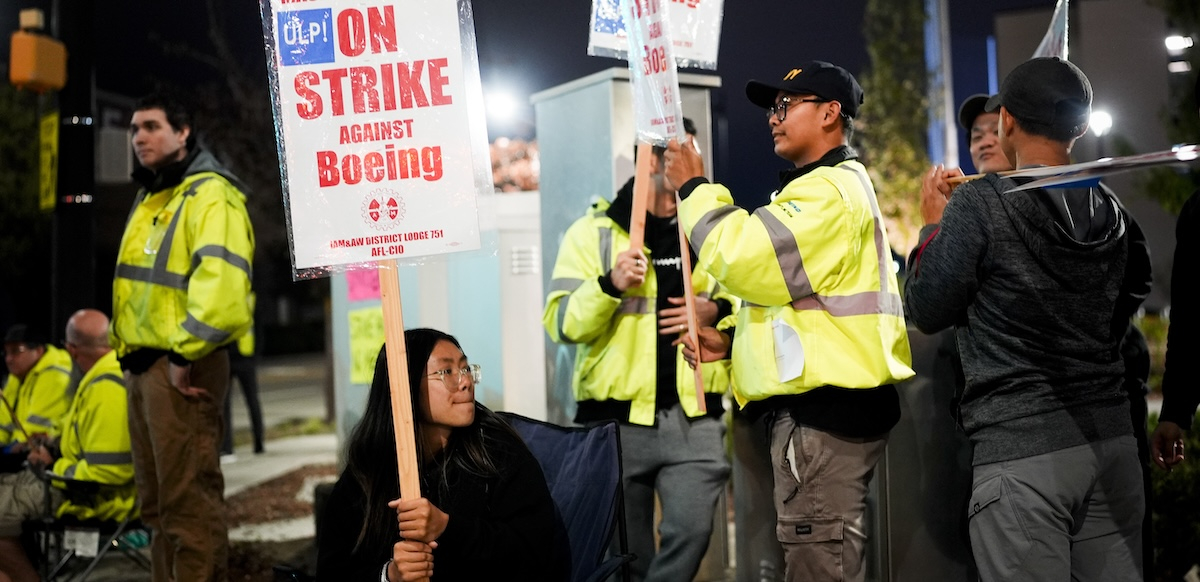 a young asian woman grips a sign that says on On Strike at Boeing. Others around her have signs. It’s nighttime.