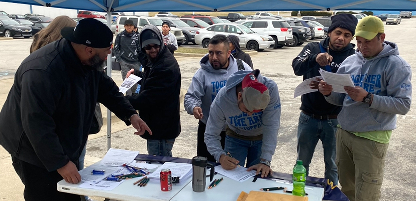 A group of men in sweatshirts gather around a table and sign things, a parking lot is behind them.