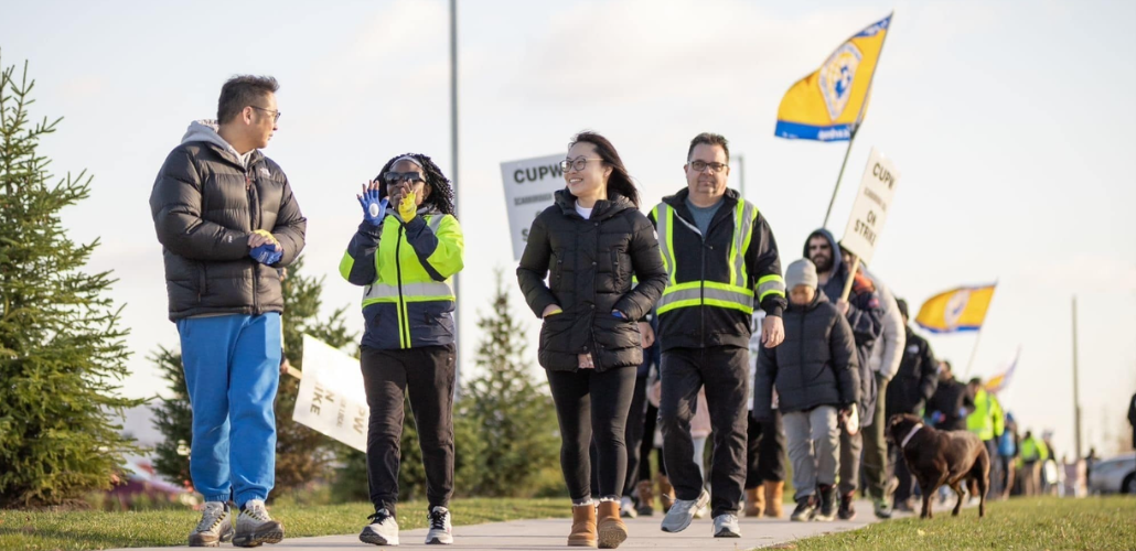 Workers silhouetted against the sky march forward in a line, smiling. Some hold "CUPW" flags aloft. The view is looking up from near the ground, giving them a heroic vibe.