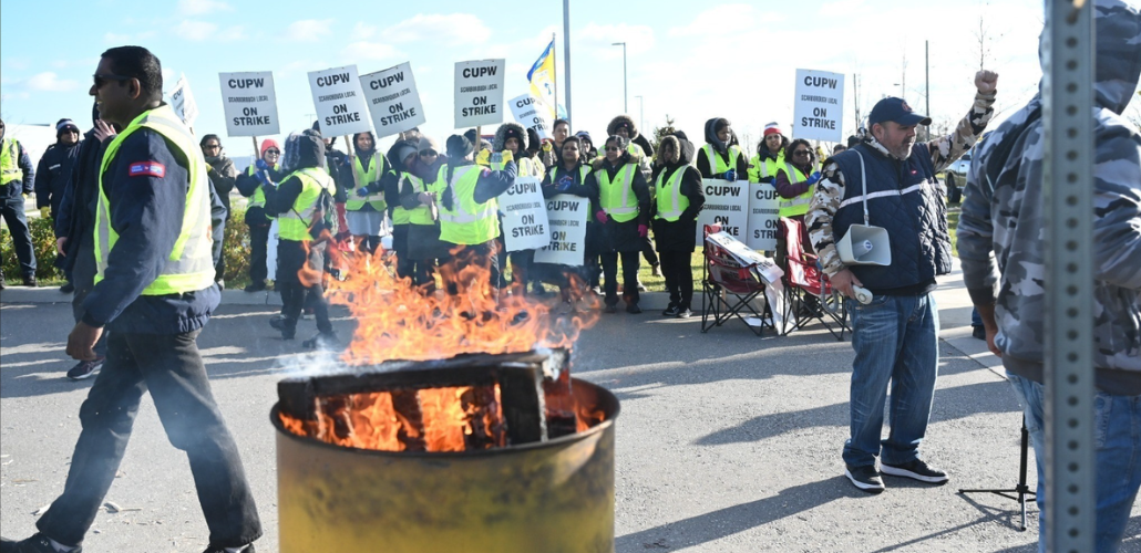 In the foreground a burn barrel, one striker walking purposefully, one raising a fist. In the background a large crowd of strikers carrying "CUPW on strike" picket signs and wearing safety-yellow vests.