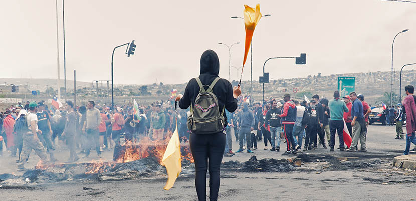 Protester holding what appears to be a flag and a torch in front of burnt out remains. Small crowd in background.