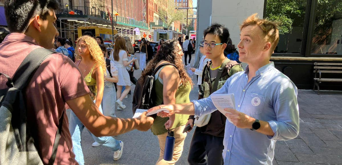 a man in a blue shirt hands a flier to a passerby wearing a backpack