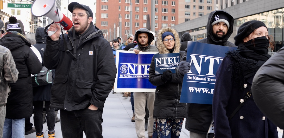 A line of men and women picketers with NTEU signs march on the right while a man with a bullhorn chants on the right