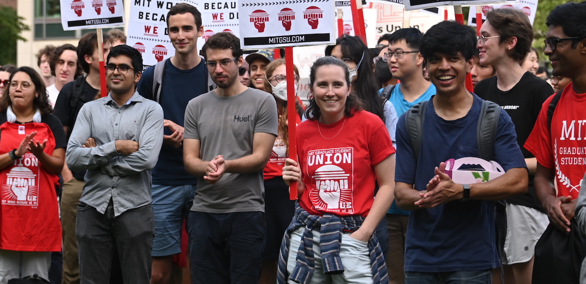 a group of 15 smiles, applauds, and holds signs saying “MIT works because WE do”