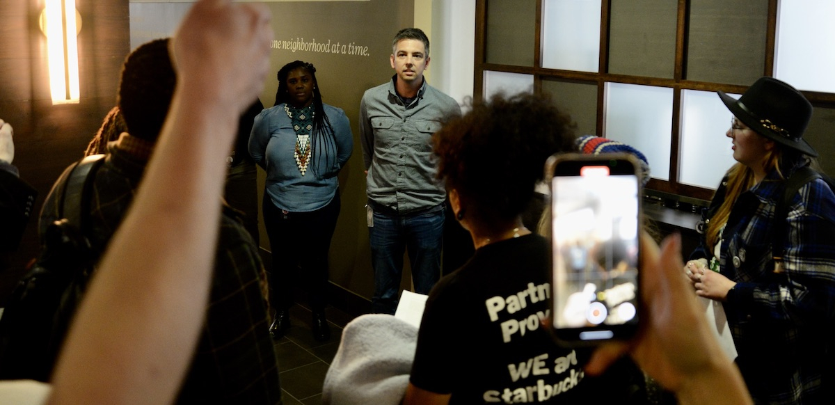 Two managers stand in a hallway as Starbucks workers gather around to confront them