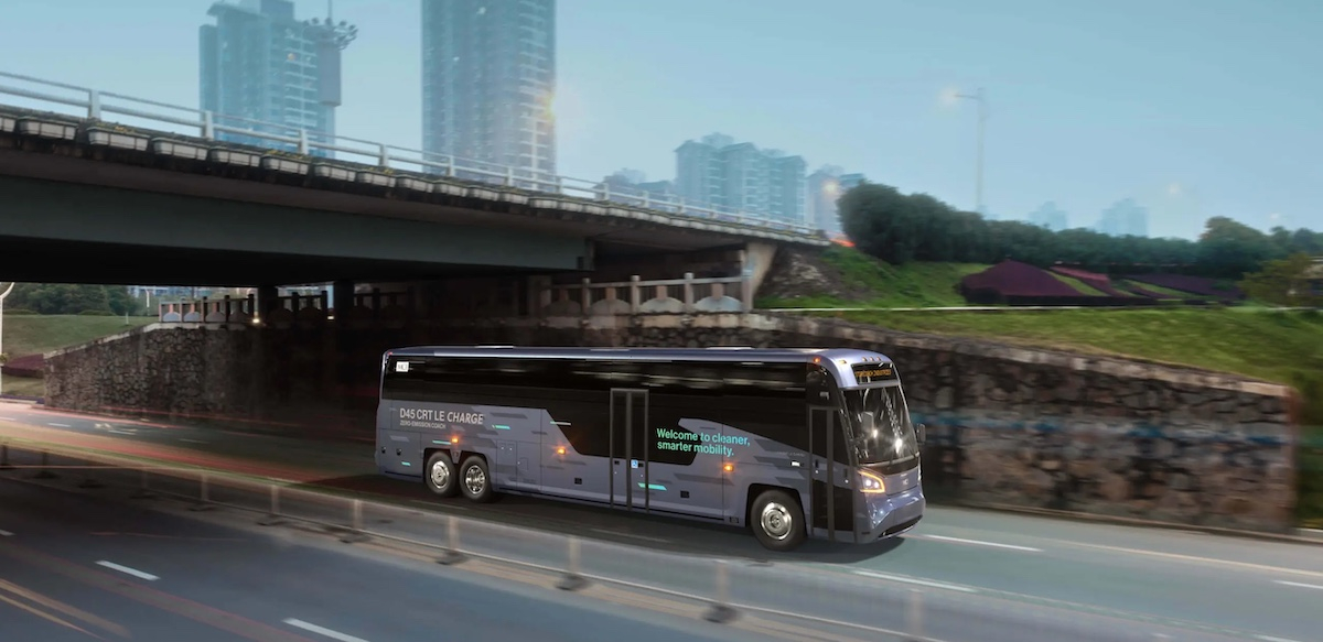 An electric coach bus drives under an underpass with city buildings in the background