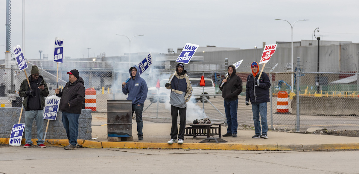 Workers six workers seen from far away hold picket signs by a burn barrel.