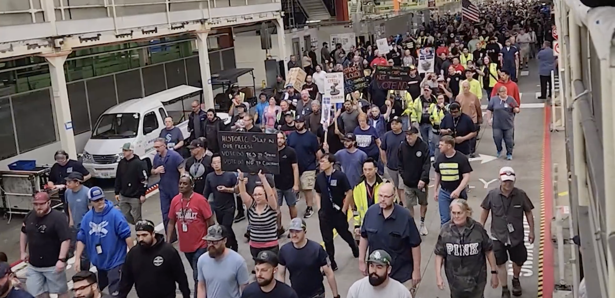 Hundreds march though an industrial plant, one sign reads: Historic Slap in Our Faces