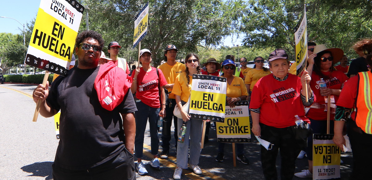 One Black man and several Asian and Latina women hold strike signs in a hotel parking lot.