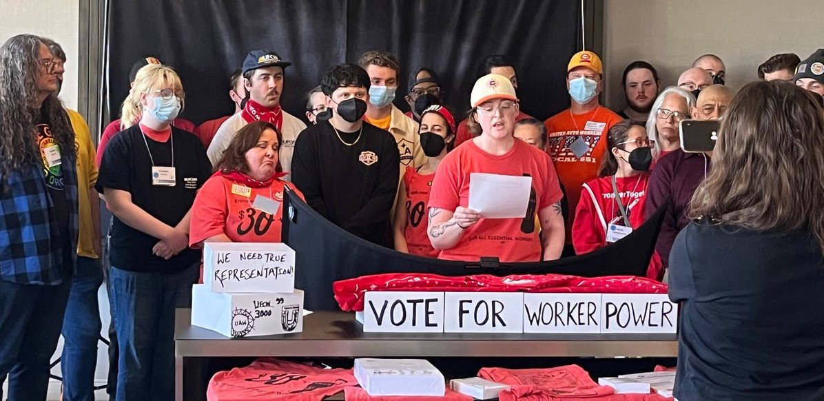 A group of serious-faced people stands indoors. Some are wearing matching red EW4D T-shirts, including Iris Scott, a white woman speaking from notes while others listen and someone in front of them holds up a phone camera to livestream the event. Some in the group wear other union shirts. Handwritten signs on the table in front of the group say "We need true reprsentation," "Vote for worker power," and "UFCW 3000, UAW, TDU" [with hand-drawn logos]. 