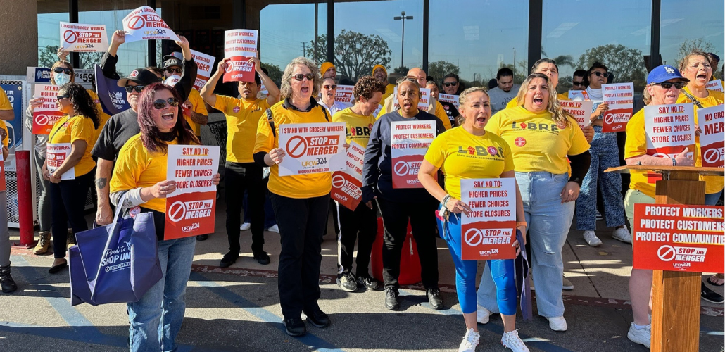 A crowd of people, mostly women, racially varied, in yellow T-shirts (some say UFCW, some say LIBRE) stand outside a grocery store, holding printed red-and-white signs, shouting together. There is a podium. Printed signs have UFCW Local 324 logo and such messages as "Stop the merger," "Say no to higher prices, fewer choices, store closures," and "Protect workers, protect customers, protect community." 