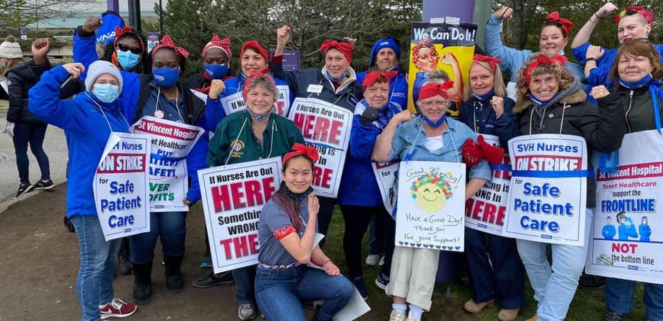 Nurses stand together holding picket signs and wearing red bandanas on their heads, a la Rosie the Riveter.