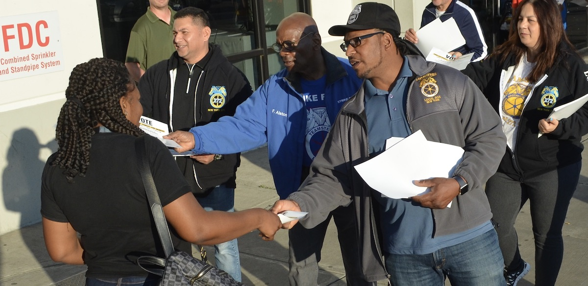 A Black man in a Teamsters 804 jacket hands a leaflet to a Black woman in a black t-shirt