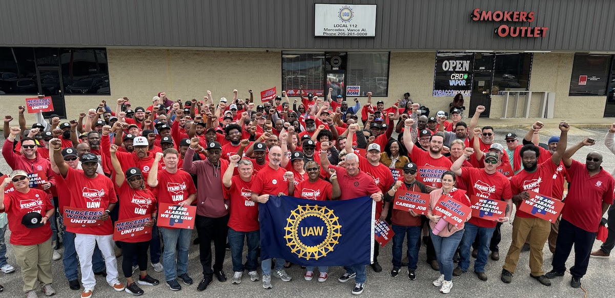 A group of a hundreds autoworkers gather outside with red shirts and fists raised.