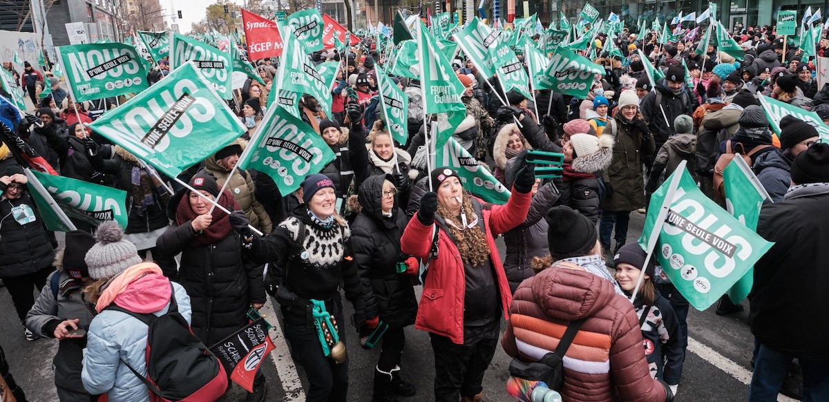 People cheer in a massive packed outdoor crowd with light green flags that say “We are of one voice” in French