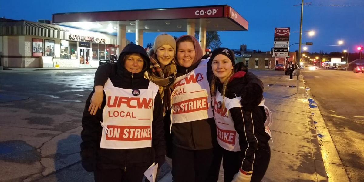 Four smiling women wearing "UFCW Local ON STRIKE" signs stand in front of a Co-op gas station, arms around each other's shoulders