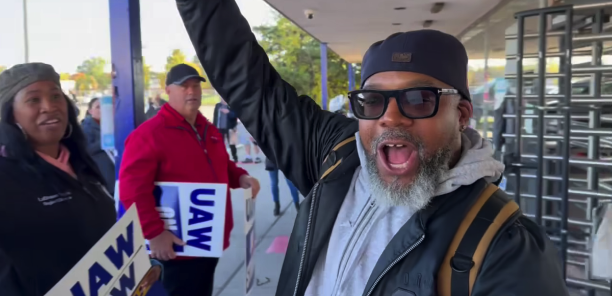 A Black man in sunglasses with an exuberant expression exits a factory turnstile with his right arm raised.