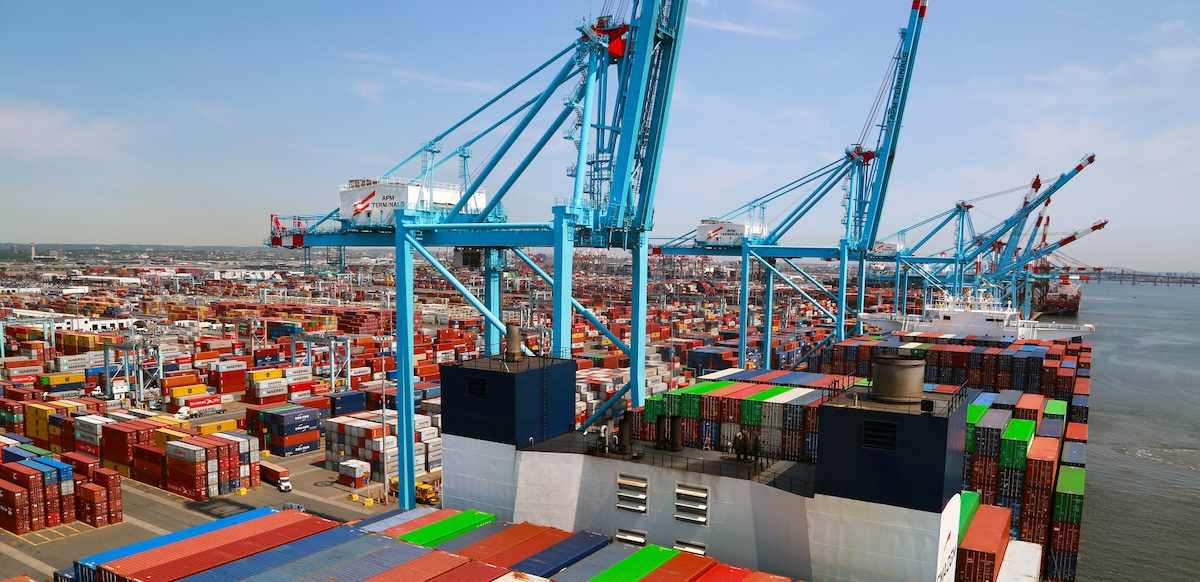 A large ship piled with containers is shown next to a dock with cranes.