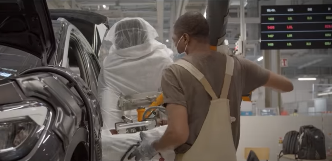 A masked worker in overalls place a seat into a car on an assembly line.