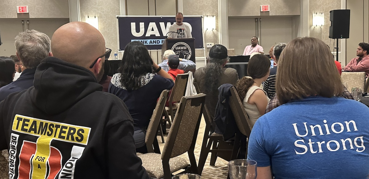 An audience listens to a man at a lectern. One audience member has a blue shirt with “Union Strong” on the back.