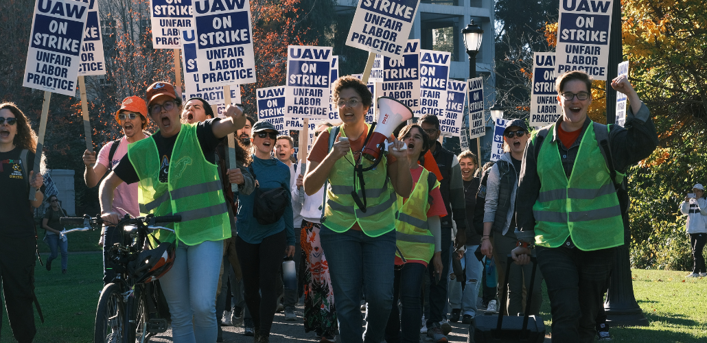 Strikers march outdoors. The three in front, wearing neon vests of picket captains, are chanting energetically, two with fists in air, one holding bullhorn. Crowd behind them holds "UAW on strike" picket signs.