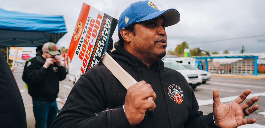 Picketer in IAFF hoodie and ballcap stands on a sidewalk, speaking and gesturing. Over his shoulder is a printed picket sign that says "End the Boeing Lockout. Boeing Firefighters - Fighting for Your Safety!"