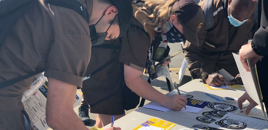 Three people in UPS uniforms and masks bend over a table of campaign literature, signing cards