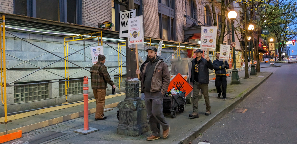 Four workers carrying picket signs walk in a circle on a downtown corner in dawn or twilight. One makes eye contact with camera.