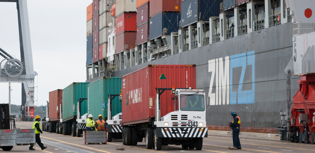 Four port workers in hardhats and neon vests look tiny as they stand beside a line of trucks with a stack of huge containers behind them.