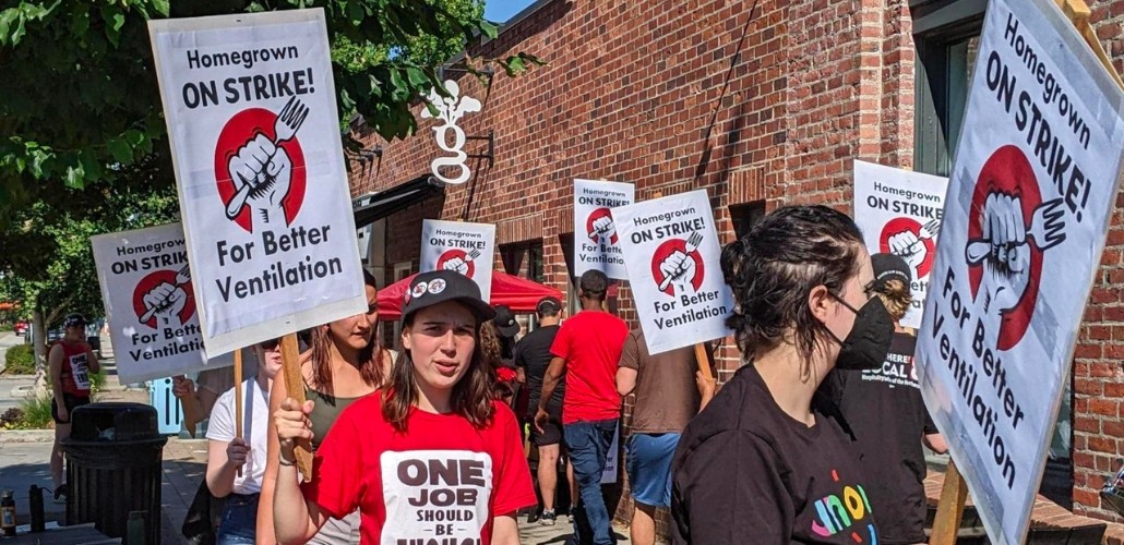 People picket in a circle outside a brick building. Their printed signs say "Homegrown on Strike! For Better Ventilation" with a logo of a fist holding a fork. Person in the foreground wears a red UNITE HERE shirt with the slogan "One job should be enough."