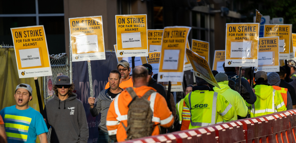 Picketers in high-visibility garb file alongside a plastic barrier. Printed signs read "ON STRIKE FOR FAIR WAGES AGAINST GLY CONSTRUCTION"