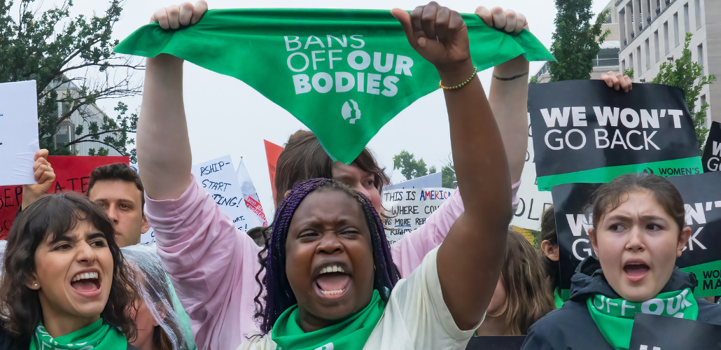 Women shout and raise their fists at a rally. Many are wearing green bandannas and one is holding the bandanna up in the air: it says "Bans off our bodies." A sign held up in background says "We won't go back."