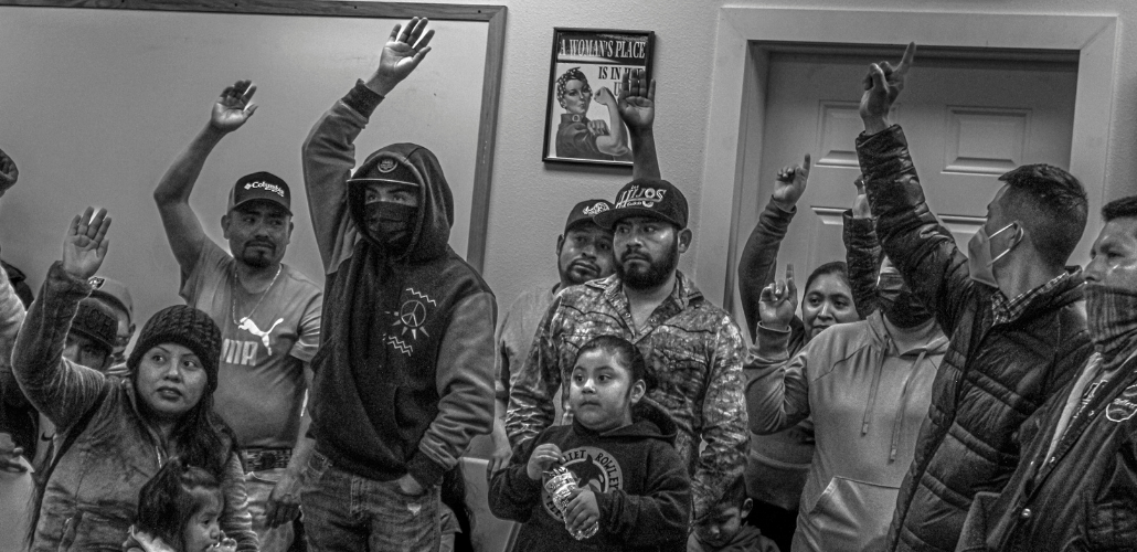 Black and white photo shows workers raising their hands, some holding kids, indoors with a small Rosie the Riveter poster on a nearby wall.