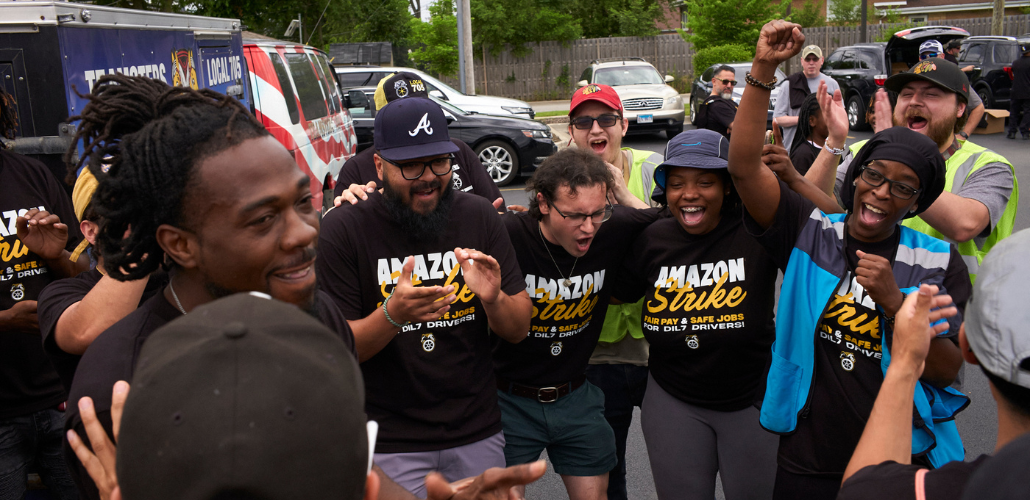 A young, racially diverse group of workers in "Amazon Strike" T-shirts huddles in a parking lot together, cheering. Some raise fists in the air. Behind them a Teamsters Local 705 truck is visible.